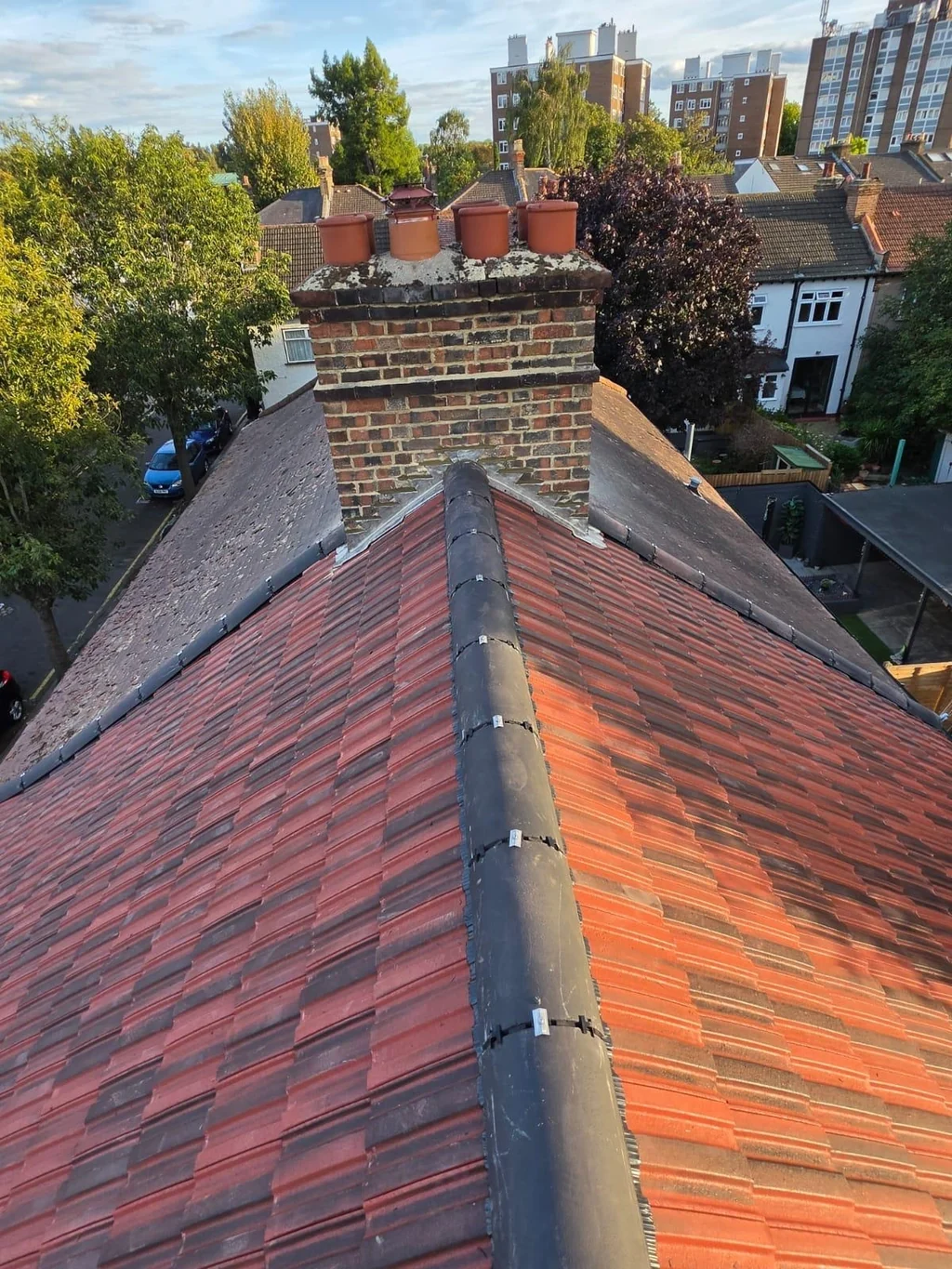 Red tiled roof and dark ridge leading to a brick chimney with urban buildings beyond
