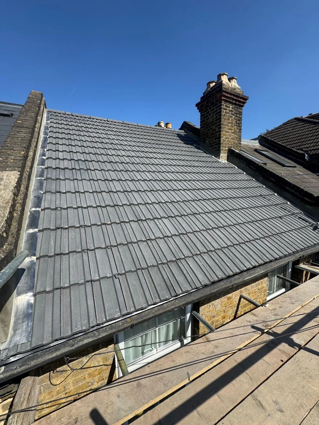 Grey tiled roof with a brick chimney and scaffold under blue sky